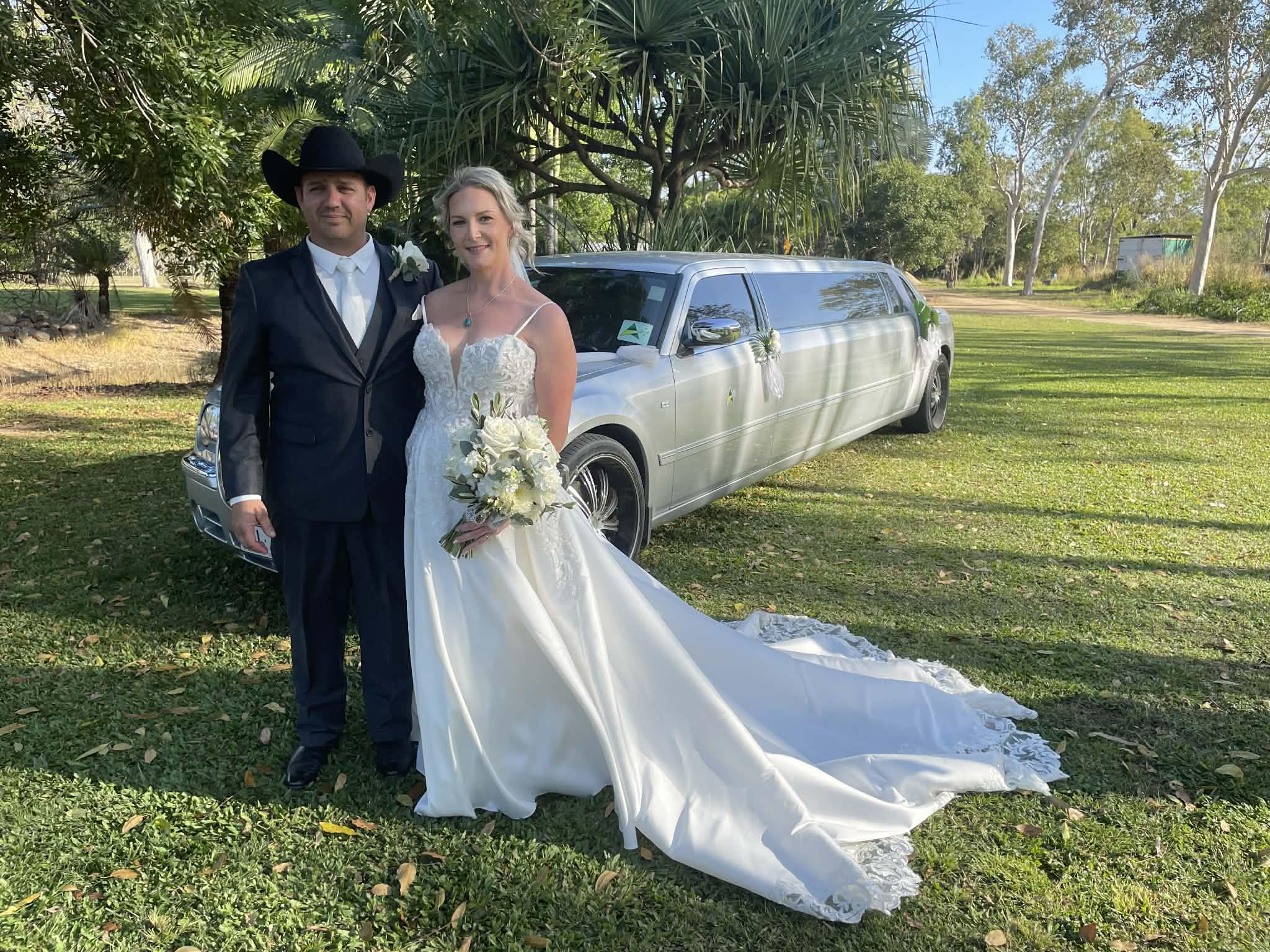 A newlywed couple, the groom in a cowboy hat and the bride in a white gown, stand in front of a white limousine on a grassy lawn.