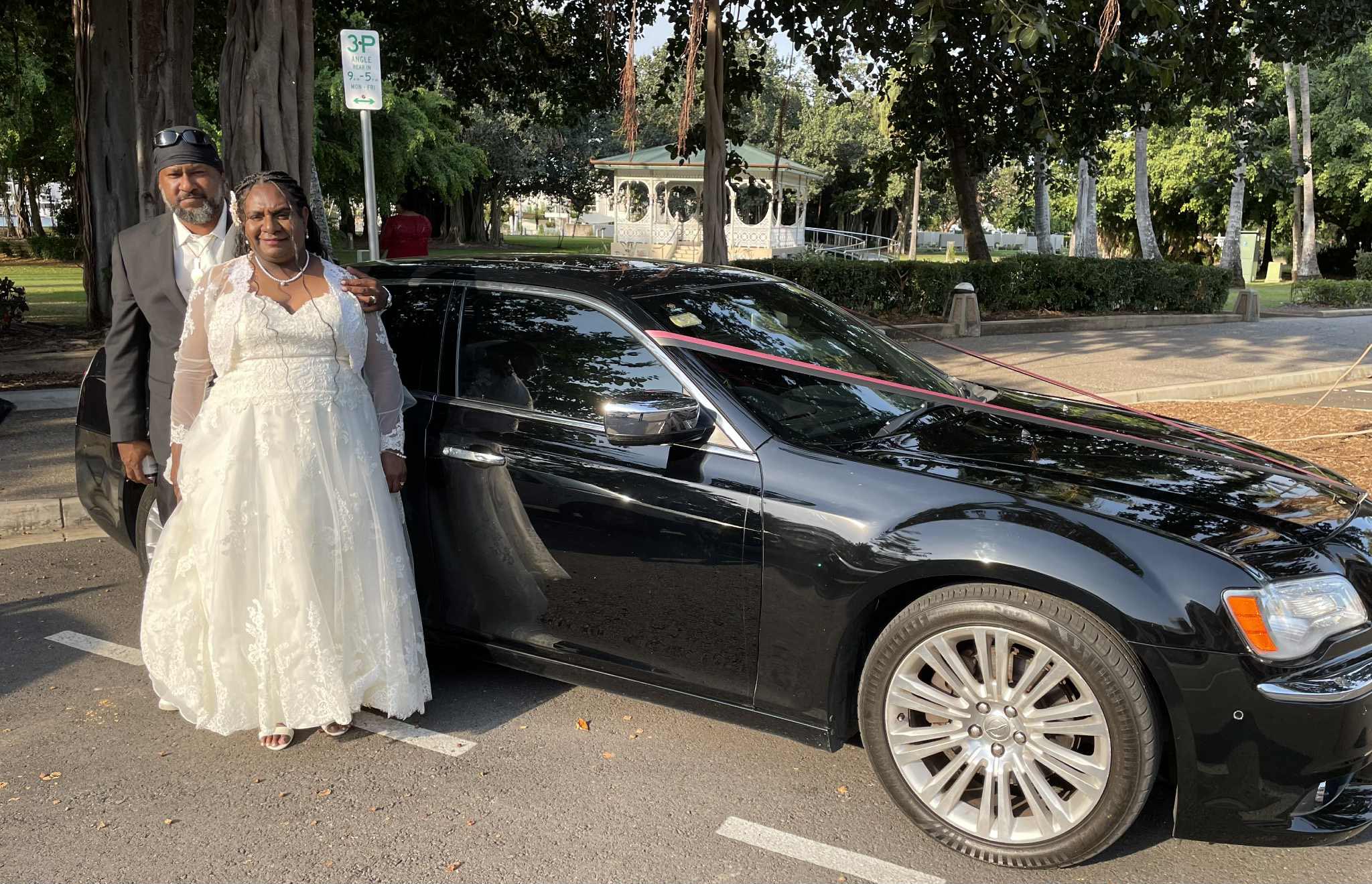 A Black couple, the bride in a white gown and groom in a dark suit with a turban, stand next to a black car in a park.
