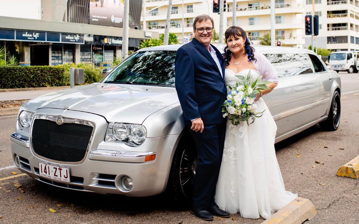 A happy bride in a white gown holding a bouquet with her groom in a blue suit, standing by a silver limousine.