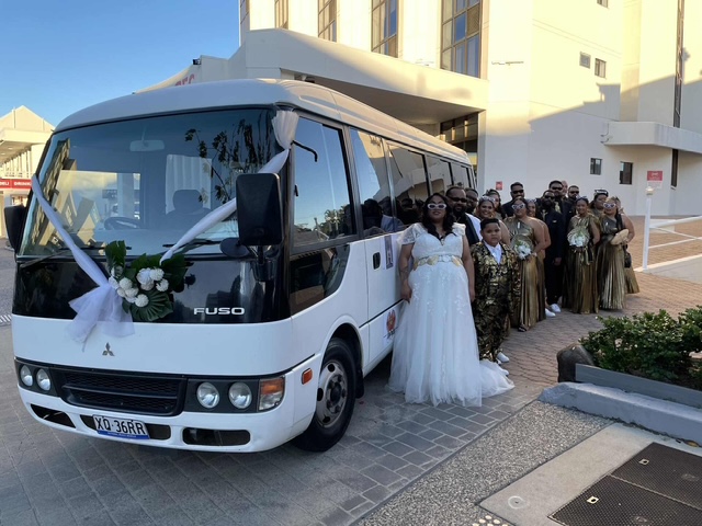 A wedding party poses by a white shuttle bus decorated with white bows and flowers, outdoors on a sunny day.