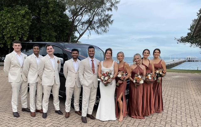 Wedding party of 10, with the bride and groom at the center, posing outdoors near a black van, with a pier and ocean in the background.
