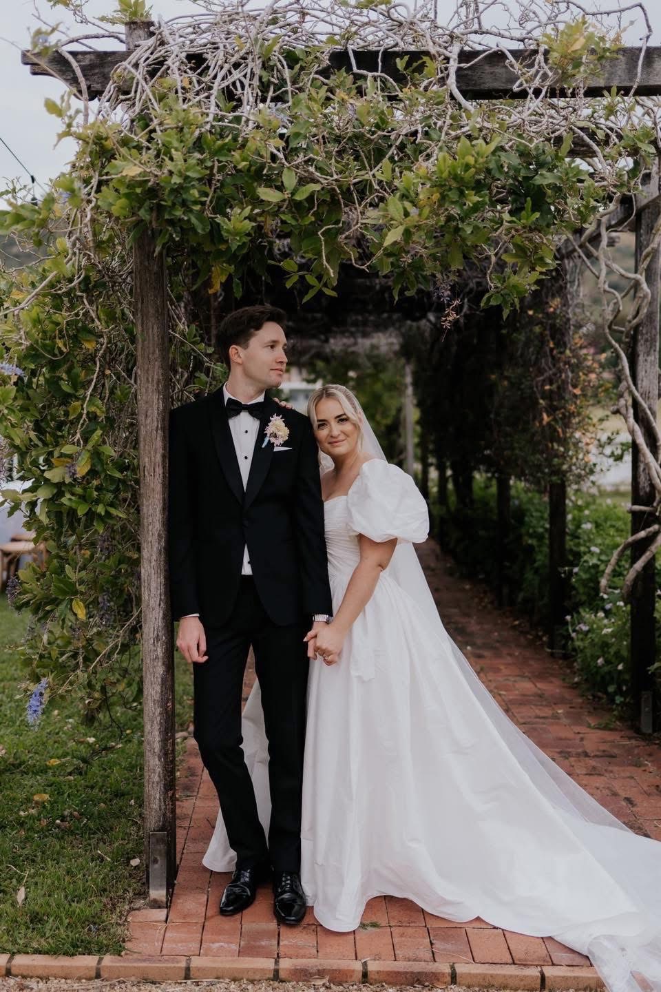A newlywed couple. The groom in a black tuxedo stands by a wooden arch covered in vines, holding hands with his bride in a white gown and veil.