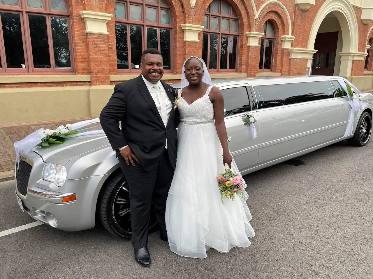 Smiling bride and groom posing next to a white stretch limousine decorated with flowers, in front of a brick building.