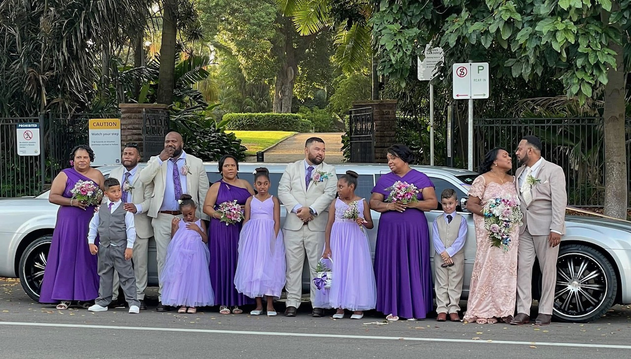 Diverse wedding party, in purple and beige attire, stands by a limousine in a lush park setting.