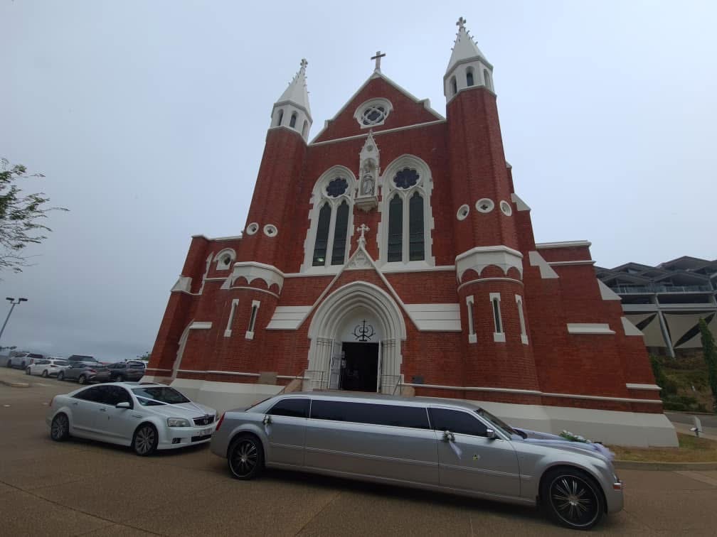 A brick church with white trim and a prominent cross on its steeple, with a silver limousine and car parked on the paved drive in front.